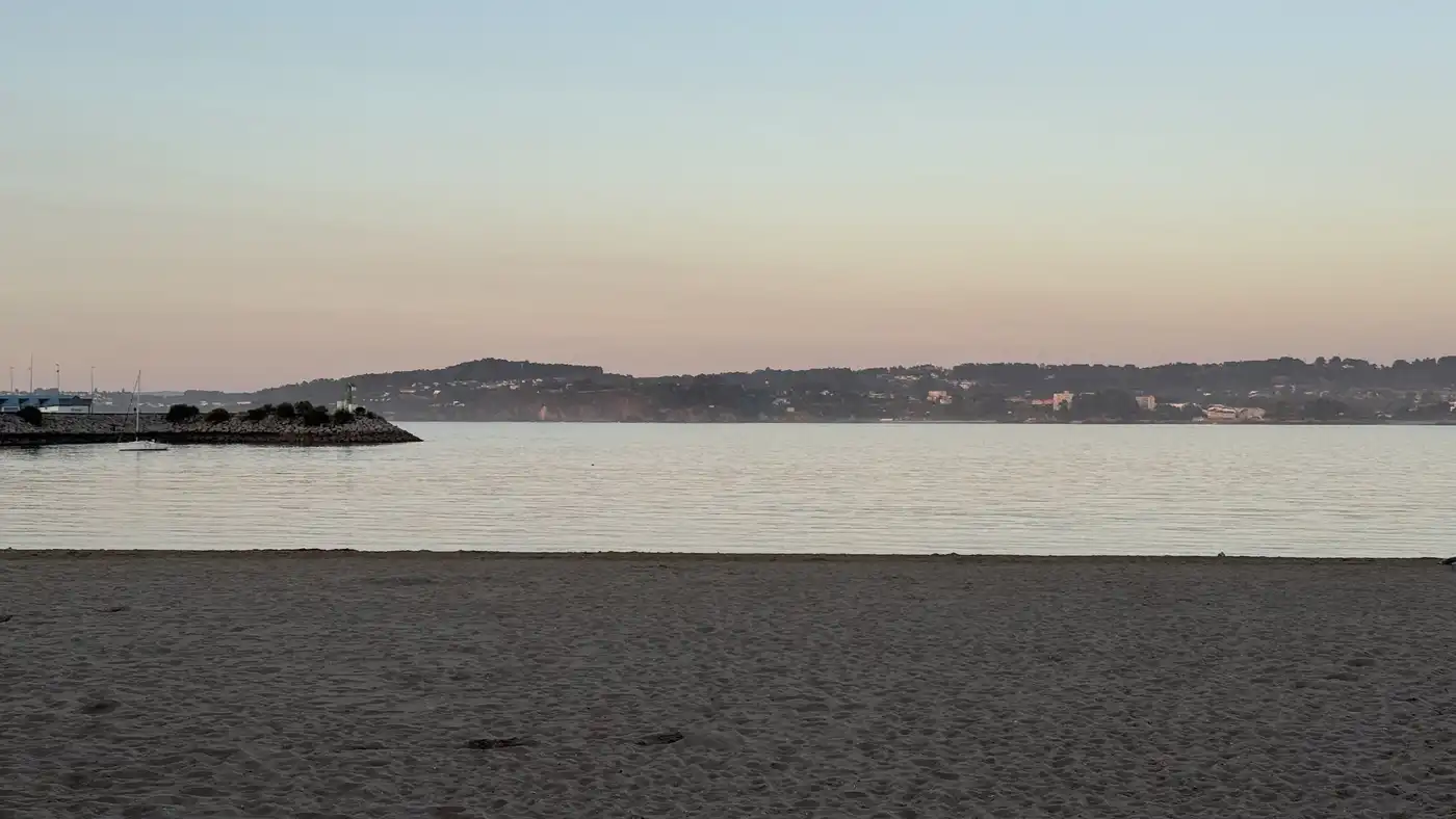 Grey sand and still water at dusk, with a low line of hills and a breakwater far off