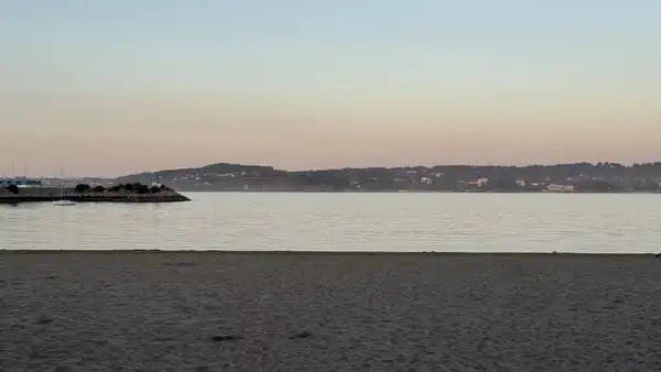 Grey sand and still water at dusk, with a low line of hills and a breakwater far off