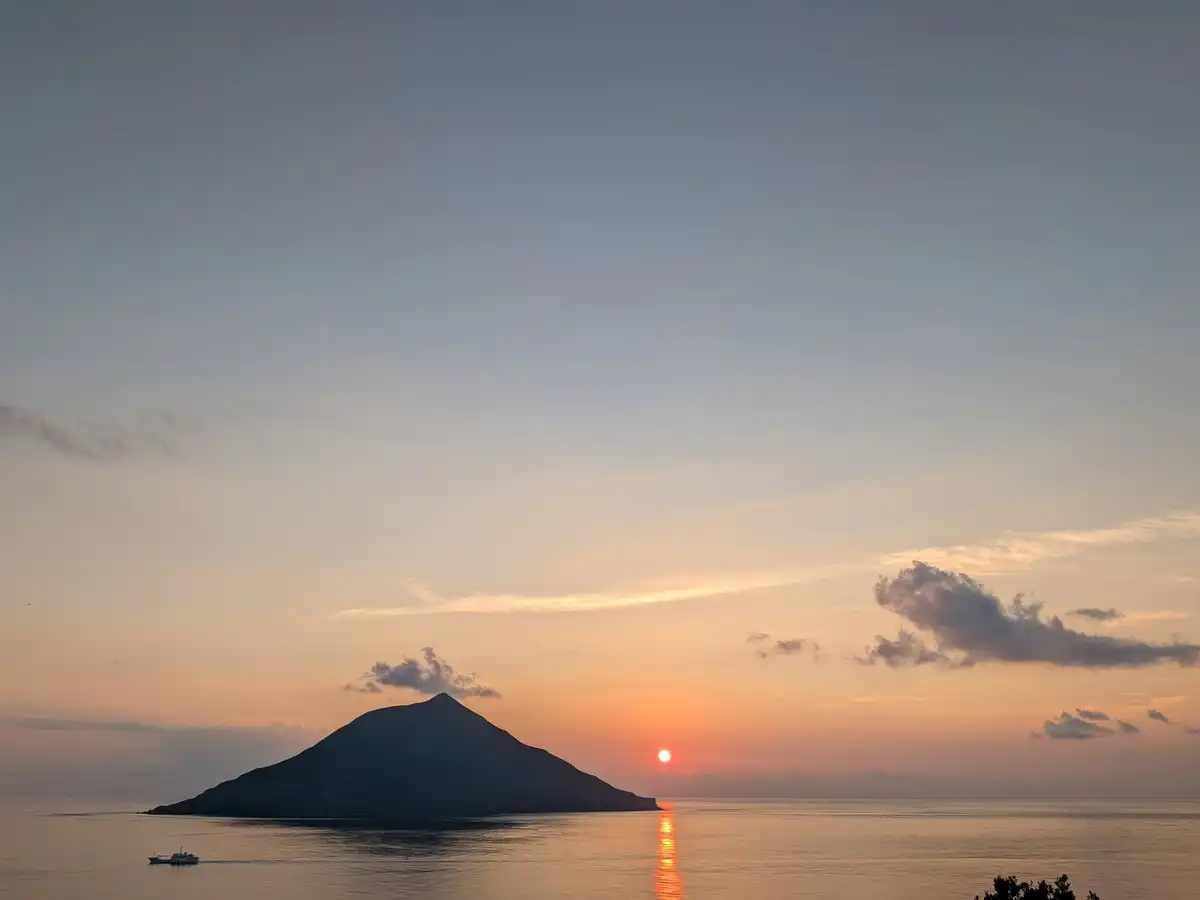 A small conical island in silhouette on a calm sea at sunset, the sun a bright point on the horizon