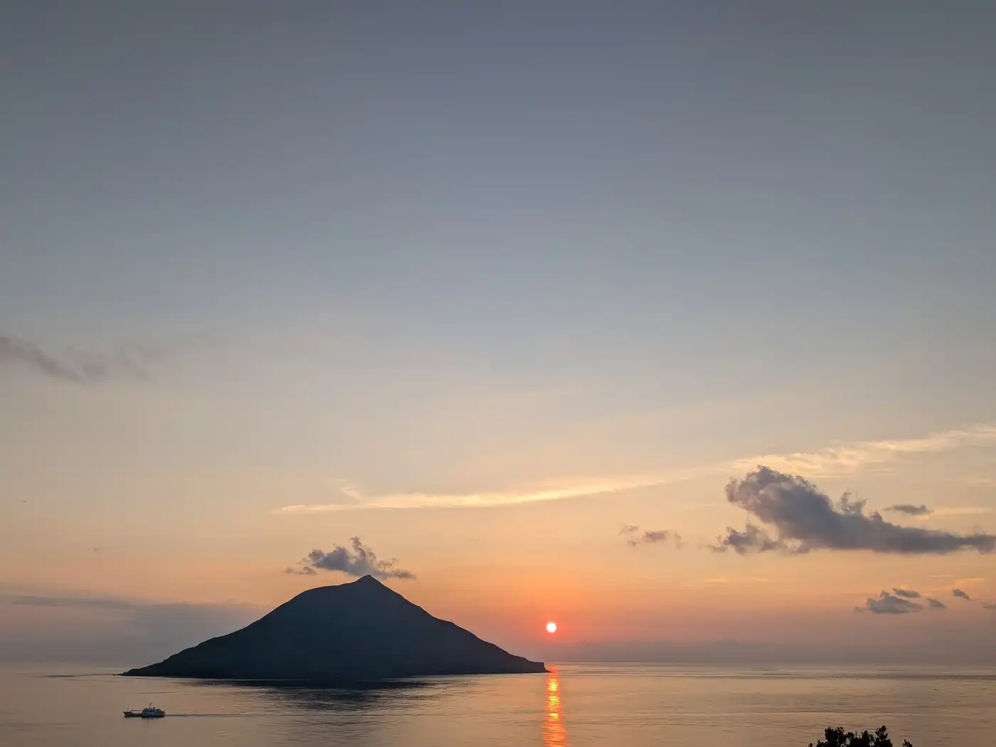 A small conical island in silhouette on a calm sea at sunset, the sun a bright point on the horizon