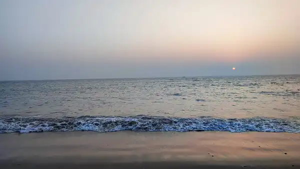 A low wave breaking on a dark beach under a pale pink evening sky with a small sun