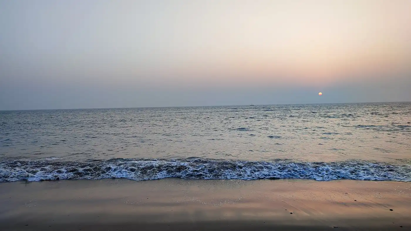 A low wave breaking on a dark beach under a pale pink evening sky with a small sun