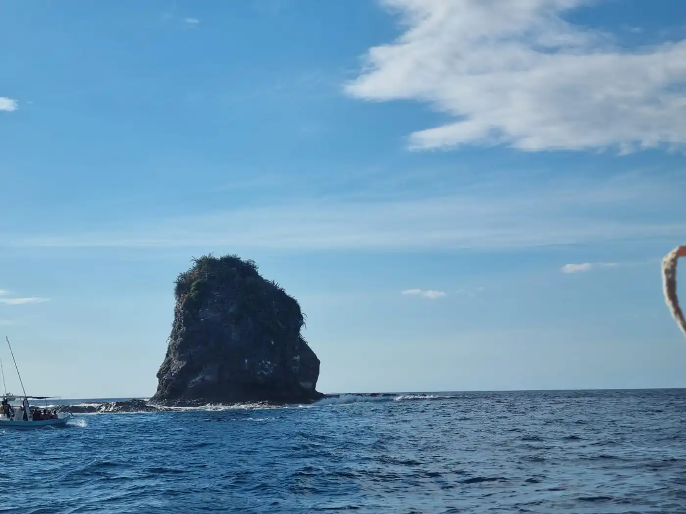 A dark rock stack rising from deep blue sea under a bright sky with white clouds