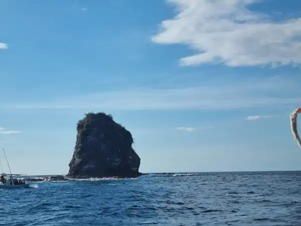 A dark rock stack rising from deep blue sea under a bright sky with white clouds